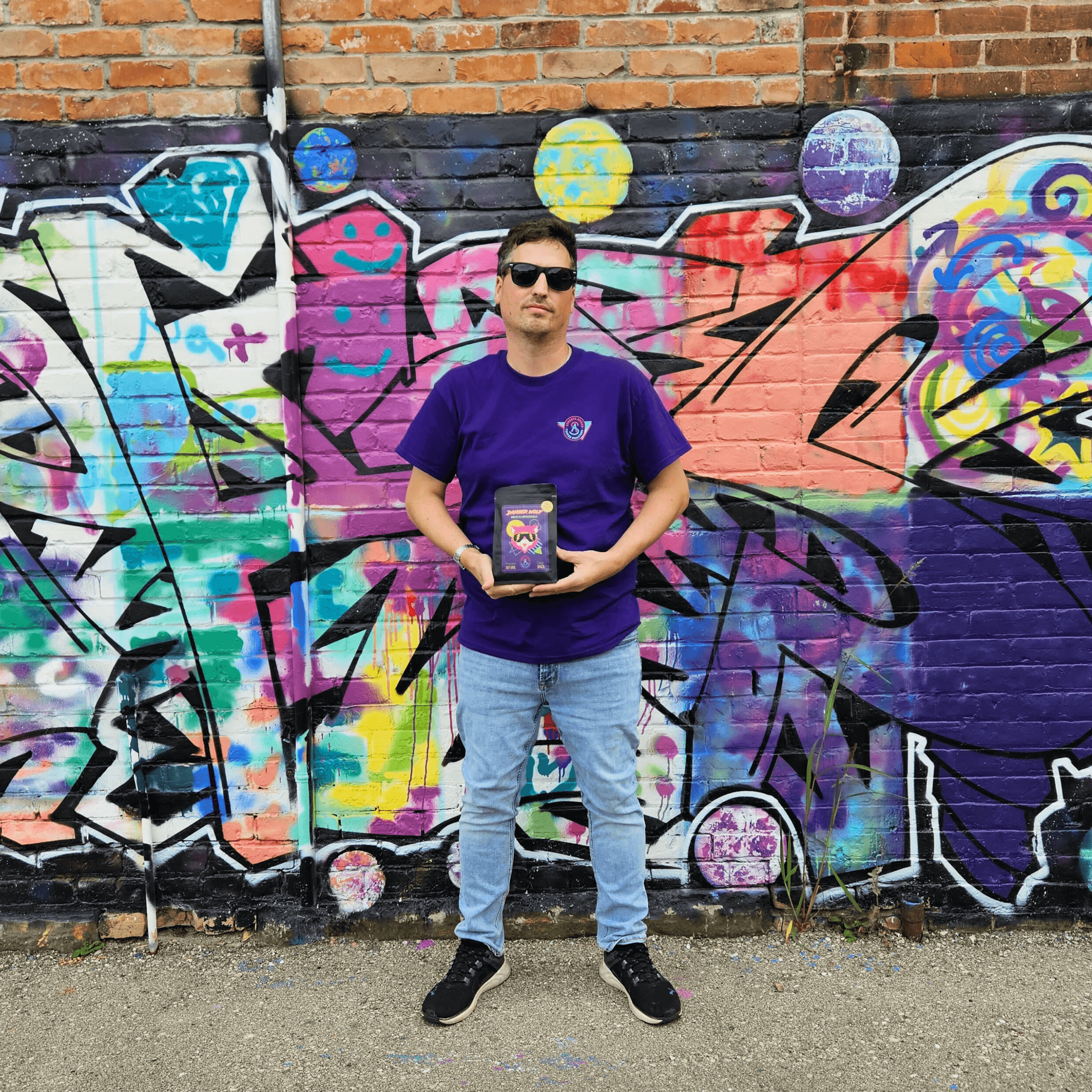 Man standing in front of a graffitied wall holding onto a bag of Danger Wolf coffee beans. The man is wearing a purple t-shirt with the Streets Ahead Coffee Roasters' logo on his left chest. 