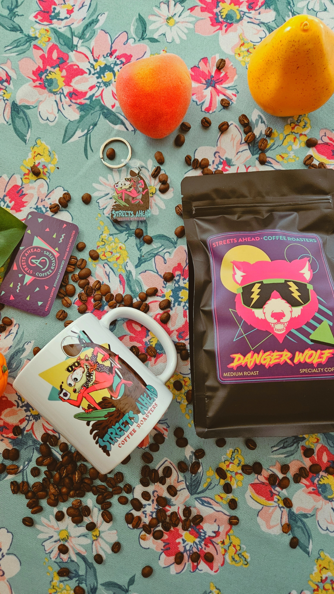 A styled flat-lay featuring a bag of coffee labeled 'Danger Wolf' and a white ceramic mug with colorful cartoon artwork, surrounded by coffee beans, fruit, and a floral fabric background.