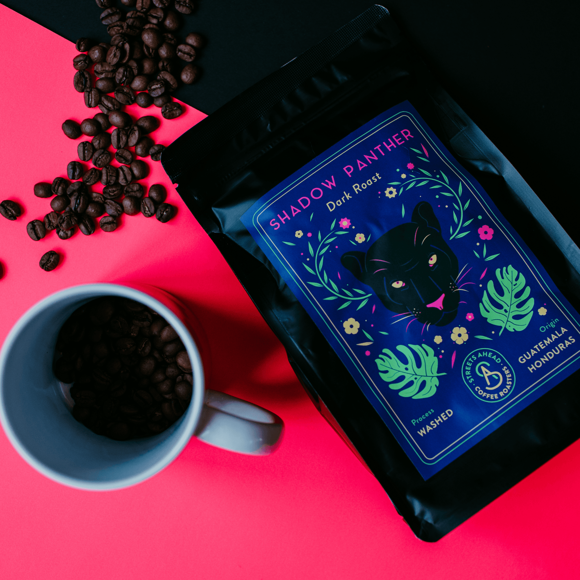 Dark roast, ground, specialty coffee bag featuring a drawing of a black panther, laying on top of a pink and black background with a coffee cup and coffee beans around it.