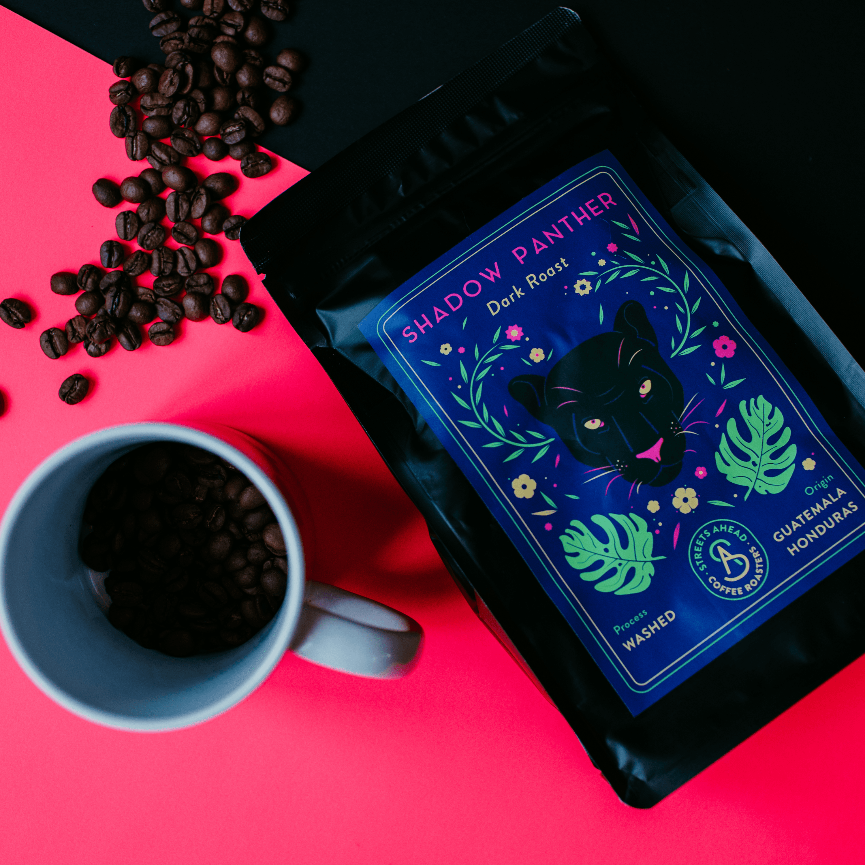 Dark roast, ground, specialty coffee bag featuring a drawing of a black panther, laying on top of a pink and black background with a coffee cup and coffee beans around it.