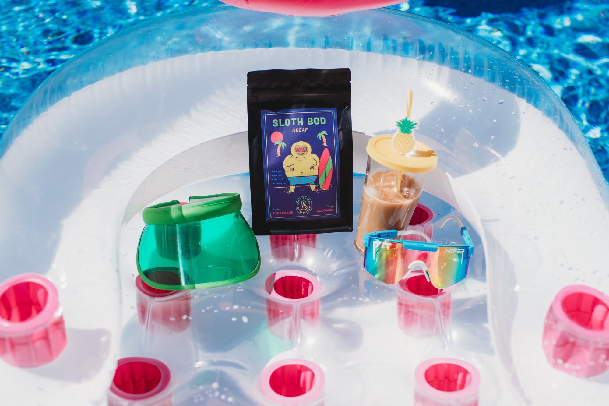 Inflatable pool float with Sloth Bod coffee package, sunglasses, and drinks on a pool background