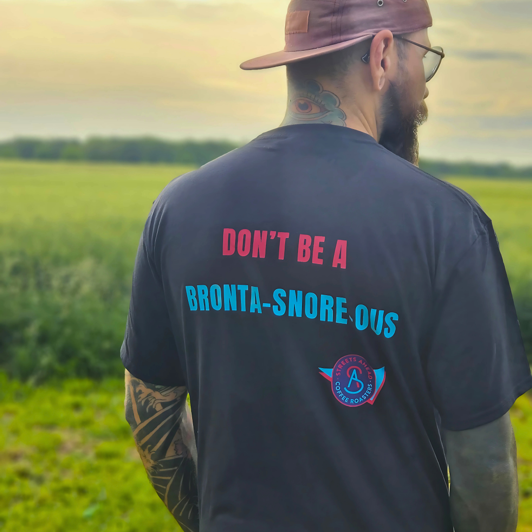 Man wearing a black t-shirt with "Don't Be A Bronta-Snore-Ous" written on the back and logo in a field