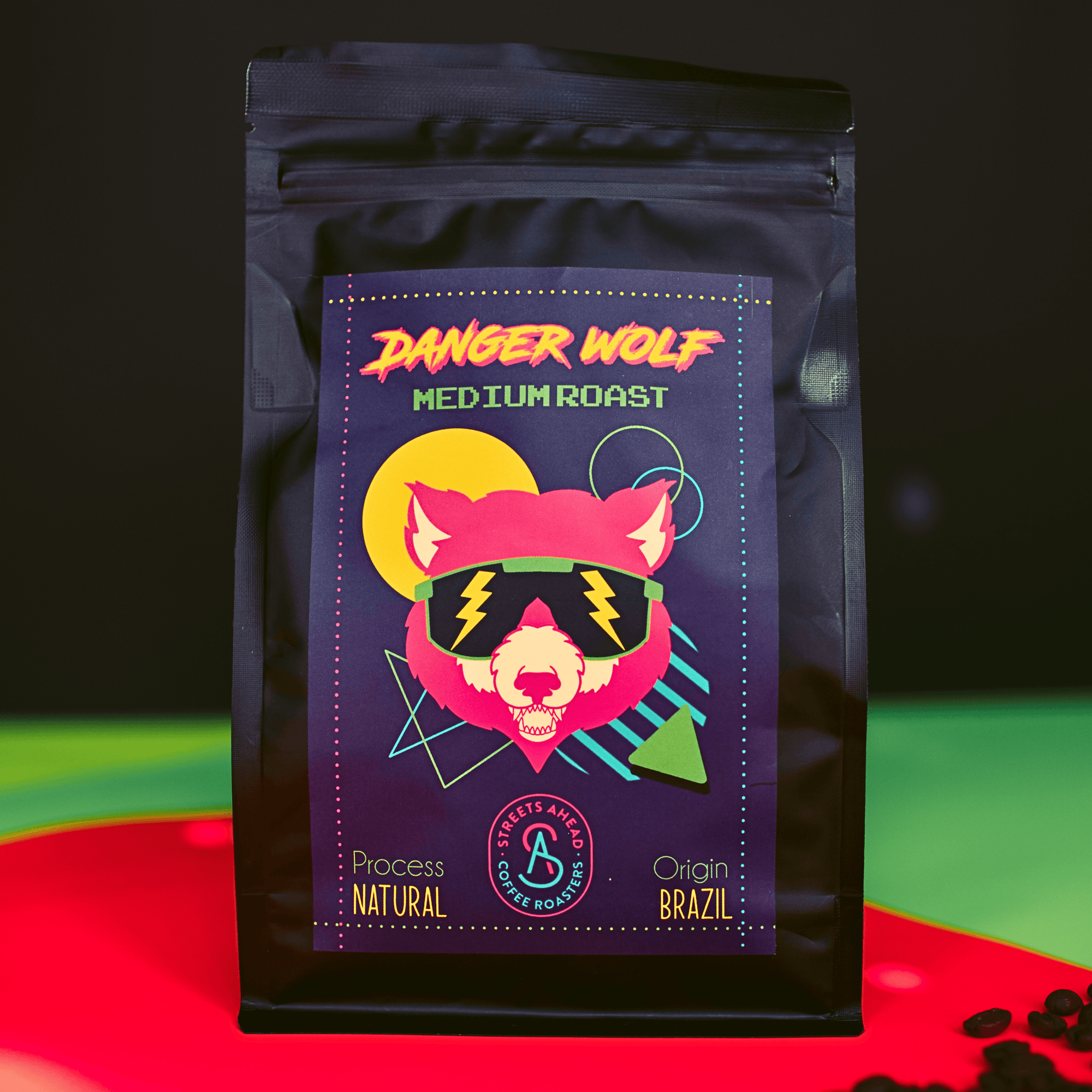 Medium roast, ground, specialty coffee bag featuring a drawing of a wolf with sunglasses, standing on top of neon paper with coffee beans surrounding it.