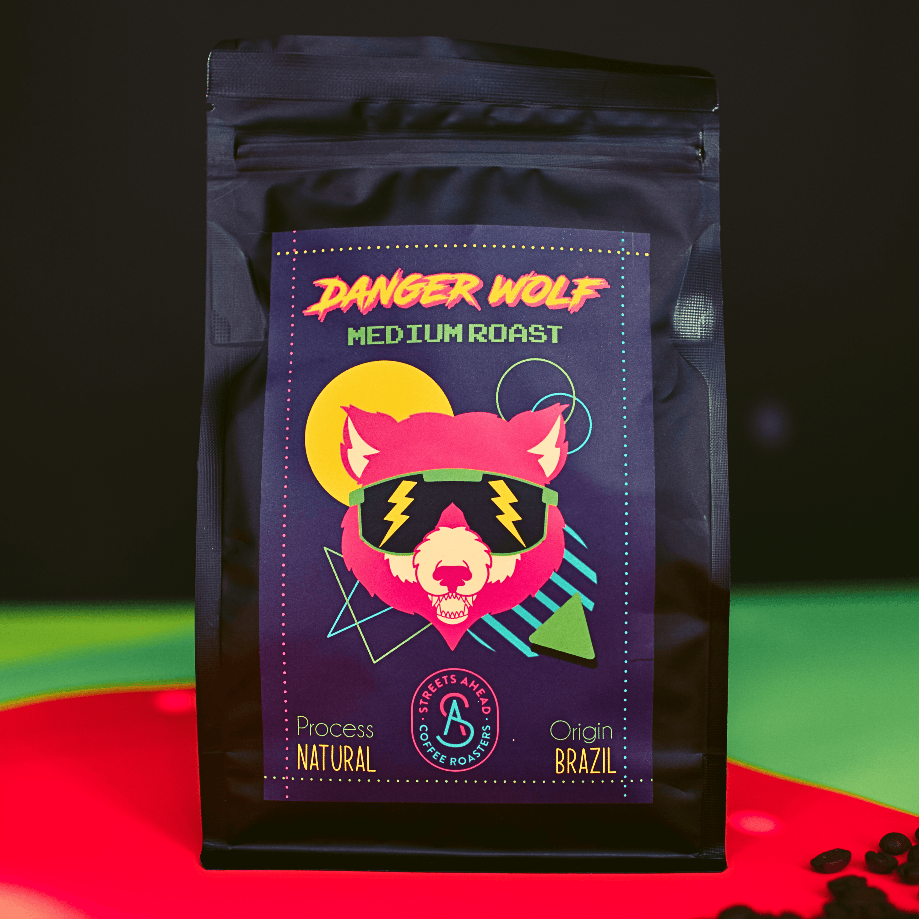 Medium roast, ground, specialty coffee bag featuring a drawing of a wolf with sunglasses, standing on top of neon paper with coffee beans surrounding it.