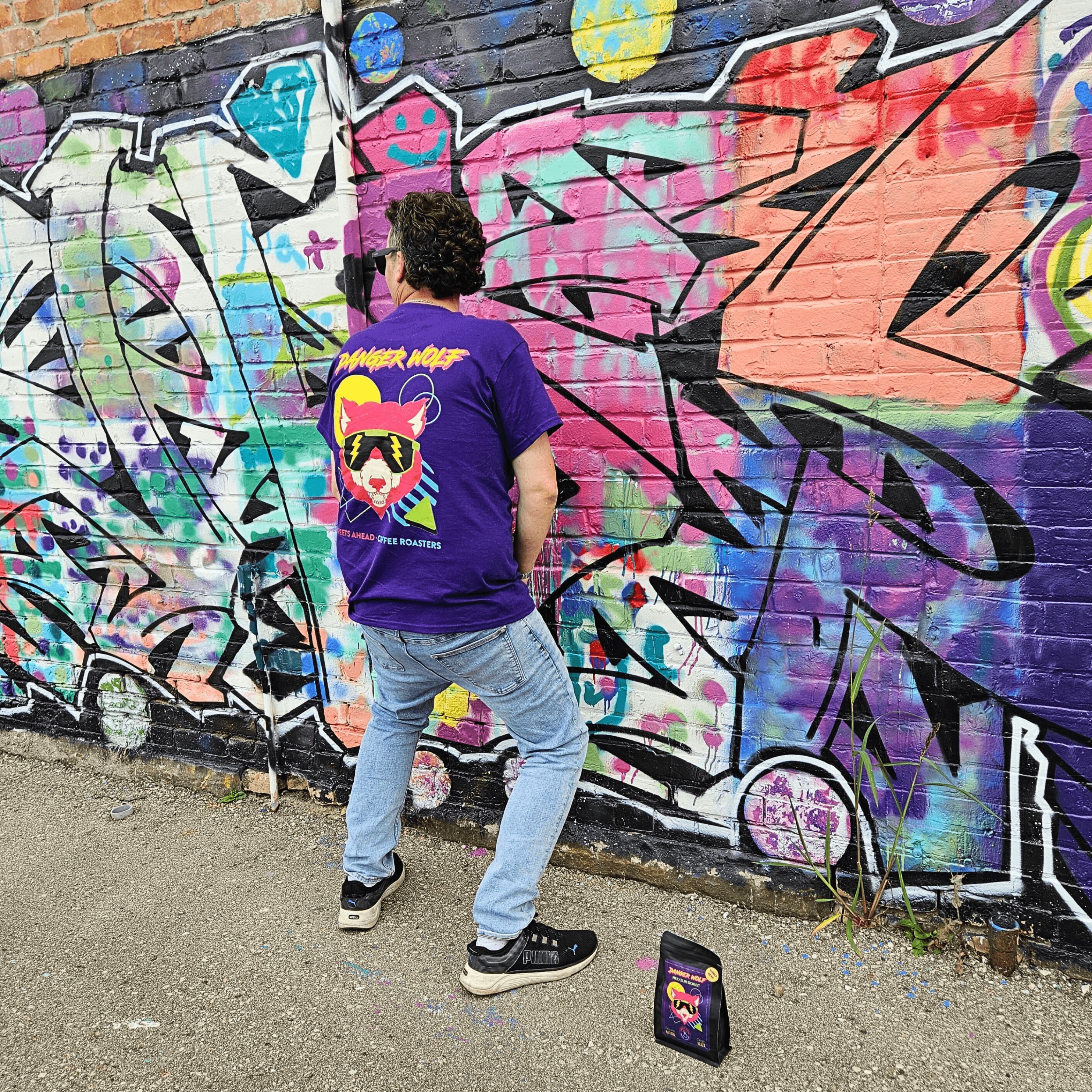 Man pretending to urinate on graffitied wall while wearing a purple t-shirt that has the Danger Wolf logo on the back. A Danger Wolf coffee bag sits on the ground beside him. 