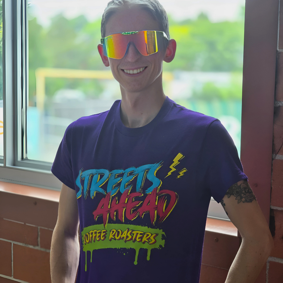 Person wearing a purple t-shirt with colorful text, standing indoors near a window.