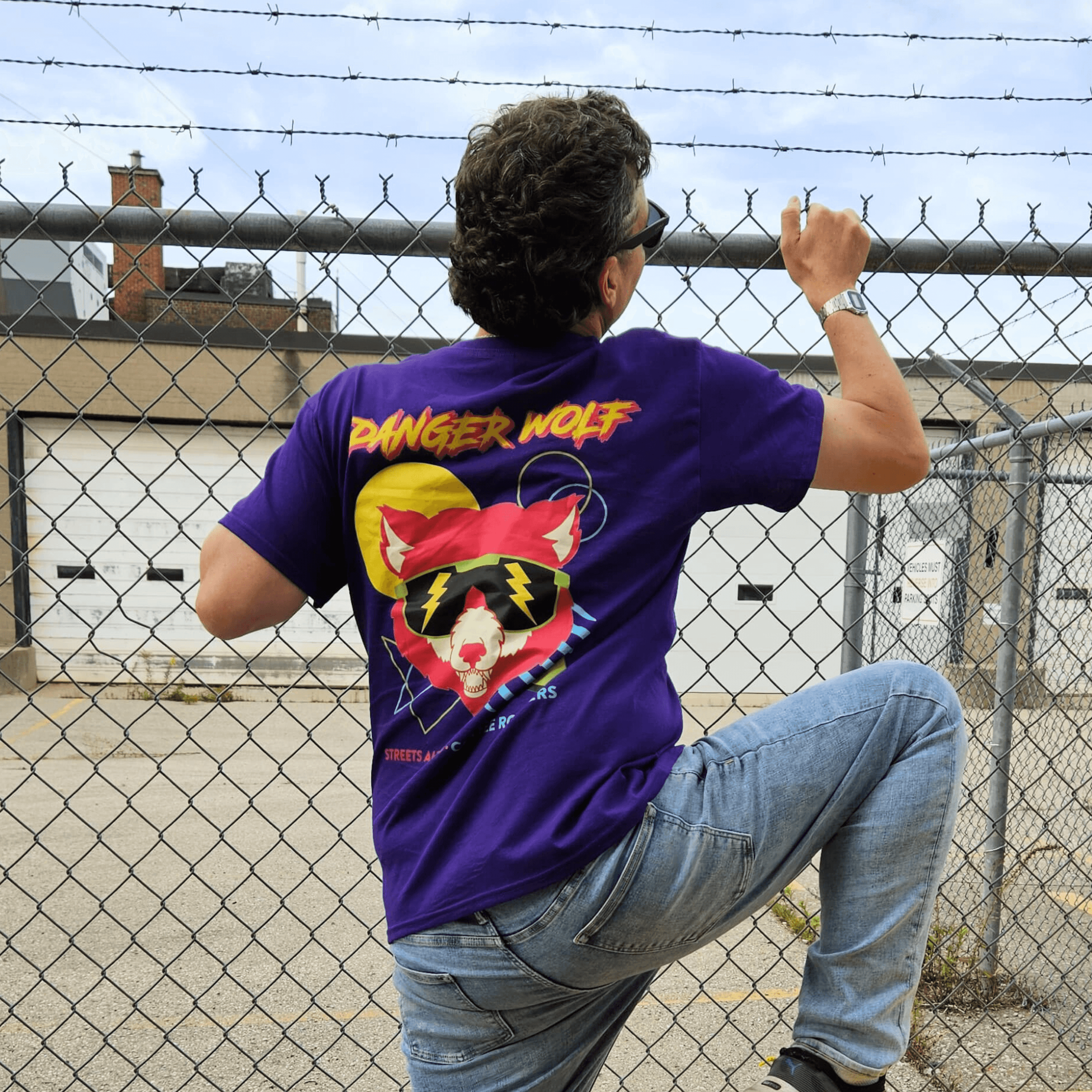 Man climbing over a fence wearing a purple t-shirt that has the words Danger Wolf on it and a design of a wolf in sunglasses on the back. 