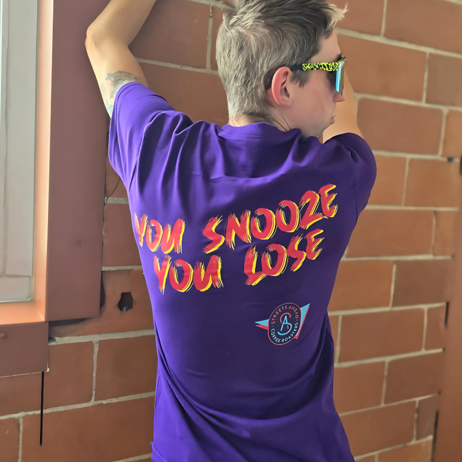 Person wearing a purple t-shirt with "You Snooze You Lose" written on the back and logo, standing against a brick wall.