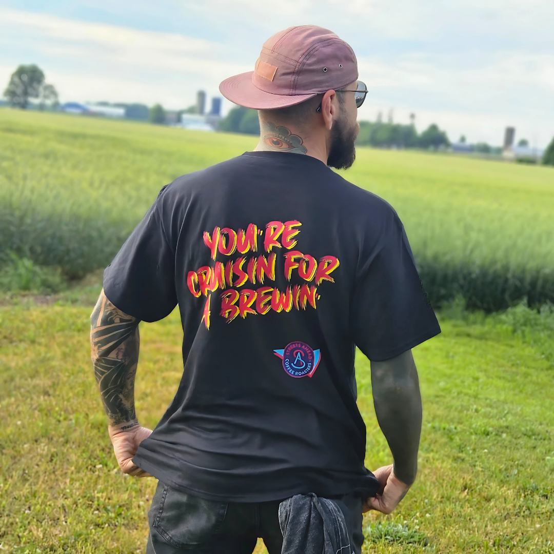 Man wearing a black t-shirt with colorful text saying "You're Cruisin for a Brewin" in an open field